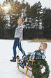 © sofiko14 - Pretty young mother pulling decorated wooden sled with her son sitting on it in winter snowy day at nature.