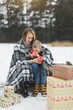 © sofiko14 - Little boy and mother sliding in the winter forest. Mom and son sitting on the sledge, covering by the checkered plaid and drinking hot drinks. Decorated present gifts around them