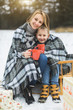 © sofiko14 - Little boy and mother sliding in the winter forest. Mom and son sitting on the sledge, covering by the checkered plaid and drinking hot drinks. Decorated present gifts around them