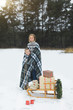 © sofiko14 - Pretty young mother with her son covered with checkered plaid, standing over the backgrounf of winter forest, pulling decorated wooden sled with Christmas wreath and present gifts on it