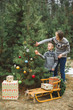 © sofiko14 - Pretty young mother and the son in gray sweaters decorating a Christmas tree in winter forest outdoors. Wooden sledge and present boxes under the tree