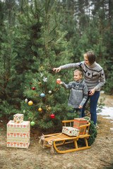 Pretty young mother and the son in gray sweaters decorating a Christmas tree in winter forest outdoors. Wooden sledge and present boxes under the tree