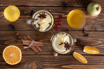 Top view of two glasses with white mulled wine with a slice of orange, lemon, apple, anise and a stick of chicken, with scattered dried fruits on a wooden table.