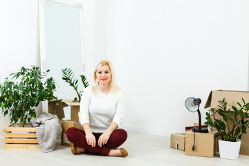 Wall Mural - Happy young woman sitting on floor near moving boxes. Young woman moving to new home. Woman looking at camera and smiling