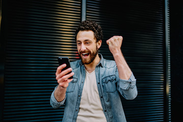 Overjoyed caucasian  bearded hipster guy excited with discount and sales in web store getting mail on smartphone, happy millennial man enjoying new modern device mobile read great news outdoors.