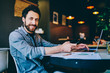 © BullRun - Cheerful hipster guy looking at camera using modern gadgets in coworking office for remote job, professional smiling male freelancer satisfied with online business sitting with laptop computer .