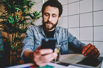 Wall Mural - Handsome caucasian hipster guy checking mail and notification on mobile phone on break in cafe, pensive male blogger share publication and content on smartphone using wifi connection in coffee shop.