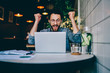 © BullRun - Overjoyed hipster guy in earphones cheers for team watching match on laptop computer during free time in cafe interior, amazed emotional male freelancer celebrating achievement and completing project.