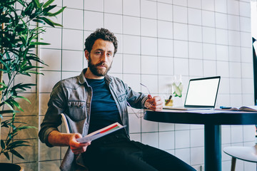 Wall Mural - Portrait of bearded intelligent hipster guy holding financial reports in paper document and looking at camera during working at digital netbook.Young man freelancer sitting at laptop with blank screen