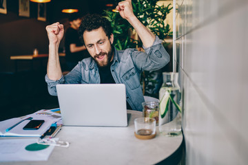 Wall Mural - Overjoyed young caucasian 20s man raising hands celebrating victory in online games on laptop computer, prosperous male freelancer excited with completed his online project successfully in cafe.