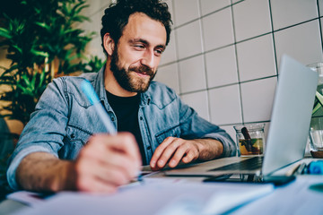 Cheerful bearded young man watching webinar online on website and writing down text notes for studying project sitting at computer in coworking.Hipster student watching tutorial and making notes