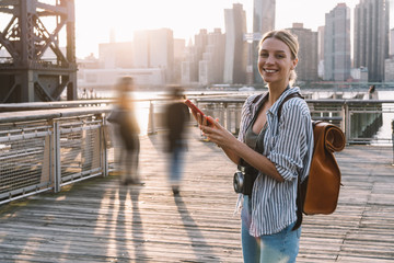 Wall Mural - Half length portrait of cheerful woman tourist with instant camera on neck and cellphone device in hands standing on American urban setting and laughing feeling excited from time for visit USA