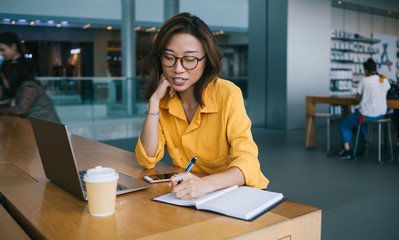 Attractive Chinese female student in optical glasses for vision correction writing information in notepad for education spending time for research, e learning with modern laptop computer concept