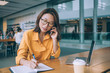 © BullRun - Beautiful Japanese female journalist receiving operator cell call for consultancy making memo notes in textbook during remote work, Asian skilled freelancer making smartphone conversation indoors