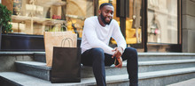 Portrait Of Afro American Male Hipster Resting After Shopping On Weekend Sitting On Stairs With Paper Bags, Smiling Dark Skinned Guy Looking At Camera Enjoying Recreation After Buying Purchases
