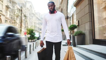 Portrait of handsome afro american guy carrying paper bags walking on city street after shopping, dark skinned male in long sleeve shirt with copy space for brand name or label strolling outdoors