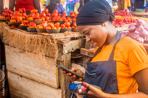 Fotomural young african woman selling in a local african market holding a mobile point of