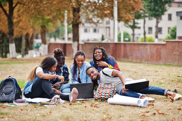 Wall Mural - Group of five african college students spending time together on campus at university yard. Black afro friends sitting on grass and studying with laptops.