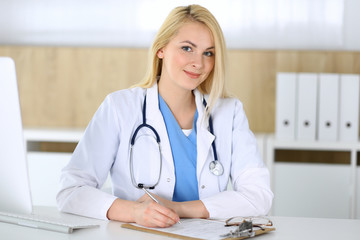 Doctor woman at work while sitting at the desk in hospital or clinic. Blonde cheerful physician filling up medical records form. Data and best service in medicine and healthcare