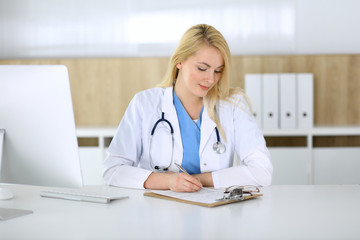 Doctor woman at work while sitting at the desk in hospital or clinic. Blonde cheerful physician filling up medical records form. Data and best service in medicine and healthcare