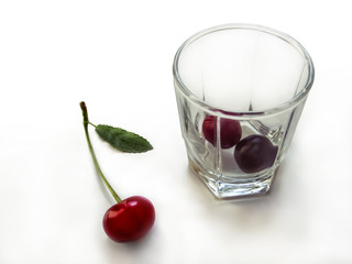 Red cherry fruit with a green stem and leaf lies near a transparent glass with two berries inside. Concept of organic drink and healthy food, isolated on white background