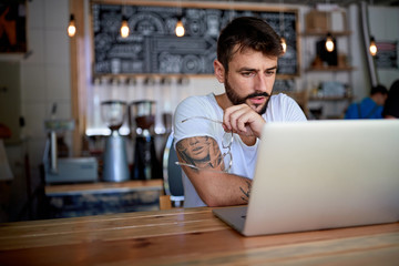 Young bearded tattooed guy in white t-shirt sitting behind the table in bar