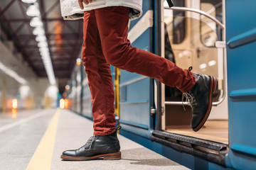 Attractive man in black shoes, burgundy pants and white jacket gets out of subway wagon. Male arrived on the last train, the platform without people. Public transport concept. Selective focus.