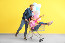 Young Couple With Shopping Cart And Balloons Near Color Wall