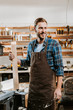 © LIGHTFIELD STUDIOS - happy carpenter in apron standing and holding wooden plank in workshop