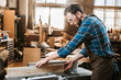 © LIGHTFIELD STUDIOS - side view of bearded woodworker in apron holding plank near circular saw