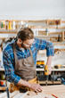© LIGHTFIELD STUDIOS - selective focus of woodworker in safety glasses and apron holding hammer drill near wooden planks