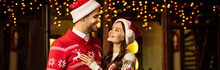 Panoramic Shot Of Happy Young Couple In Warm Sweaters And Santa Hats Smiling While Looking At Each Other