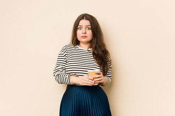 Sticker - Young curvy woman holding a coffee shrugs shoulders and open eyes confused.