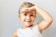 Body language. Horizontal shot of cute cheerful little girl with gathered fair hair holding palm on her forehead as if looking into distance, trying to see something far away, smiling happily
