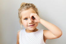 Horizontal Shot Of Adorable Pretty Girl In Beautiful Dress Posing In Studio Looking Through Binocular Made Of Her Hand, Connecting Thumb With Fore Finger. Cute Funny Female Child Having Fun, Spying