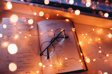 An Open Reading Book Lies On A Window With Glasses And Light From A Garland And Candles. Comfortable Reading Atmosphere