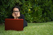 © Pajaros Volando - Woman lying down on the grass of a park watching the screen of her computer while smiling. Natural Environment. Technological concept