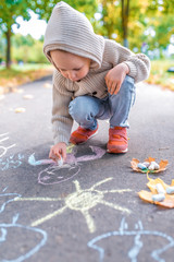 Little boy in autumn on street, draws on pavement, multicolored crayons. Warm clothes on a cold day, beige cardigan with a hood. Weekend breaks, creativity and drawings.