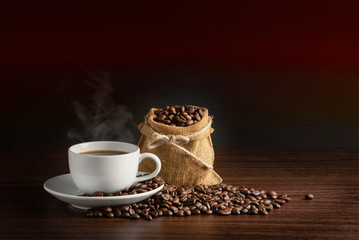 white cup of hot coffee with smoke with coffee beans and sackcloth full of coffee beans with rope on orange and black background