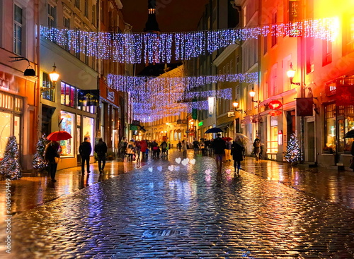 Fototapeta na szkło Deszczowa jesień Podróż do Europy w mieście Boże Narodzenie Tallinn Stare miasto ulica światło nocne ludzie spacerujący z parasolami krople deszczu odbicie refleksji na mokrym asfalcie miękkie niewyraźne światło styl życia Tilt Shift