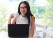 © Phuttharak - Beautiful Asian long hair business woman wearing eye glasses sitting by window with cup of coffee in her hand and computer laptop on table
