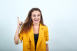 © Petro - Attractive young woman in yellow jacket pointing finger up, showing space, smiling, looking happy isolated on gray background in studio. People sincere emotions, lifestyle concept.