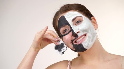 Wall Mural - Woman with clay carbo black mask on one half face and white mud on second half. Girl removing cosmetic scrab with cotton swab. Female taking care of oily complexion. Skincare