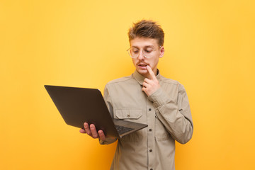 Portrait of a thoughtful young man in shirt and glasses holding a laptop in his hand and looking into the screen with a pensive face. Funny freelancer uses a laptop on a yellow background and thinks