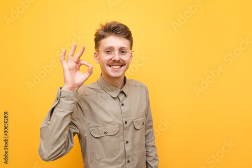 Cuadros en Lienzo Happy guy with mustache and glasses shows OK gesture and smiles against yellow background, looks into camera and wears shirt