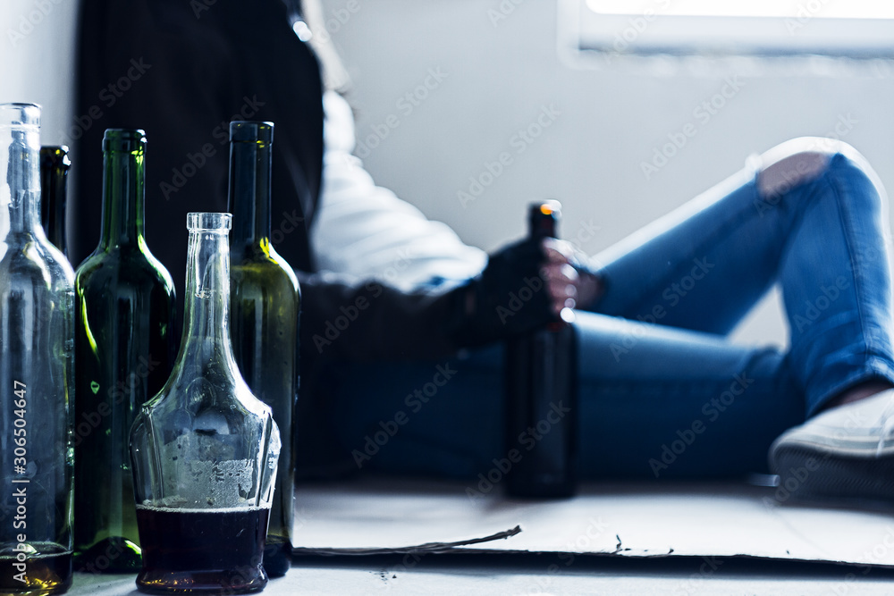 Closeup empty glass bottles. On background homeless drunk woman is sitting on cardboard on floor in abandoned house. Addicted teenage girl drunkard is drinking beer. Alcoholism, alcohol abuse concept. - obrazy, fototapety, plakaty 