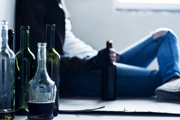 Closeup empty glass bottles. On background homeless drunk woman is sitting on cardboard on floor in abandoned house. Addicted teenage girl drunkard is drinking beer. Alcoholism, alcohol abuse concept.