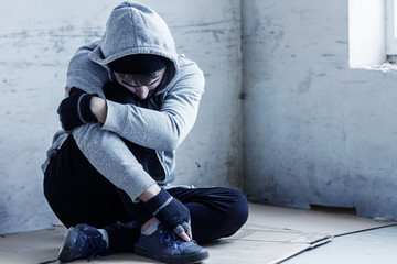 Unhappy guy in hood is sitting on cardboard on floor in abandoned building. Homeless sad man lost everything because of addiction. Social problems in society. Loneliness and depression concept.