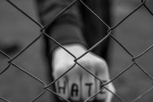 Aggressive  Teenage Boy Showing Hes Fist Behind Wired Fence At The Correctional Institute, The Word Hate Is Written On Hes Hand, Focus On The Wired Fence, Conceptual Image Of Juvenile Delinquency .