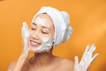 Close up beauty portrait of a laughing beautiful half naked Asian woman applying face cream and looking away isolated over orange background